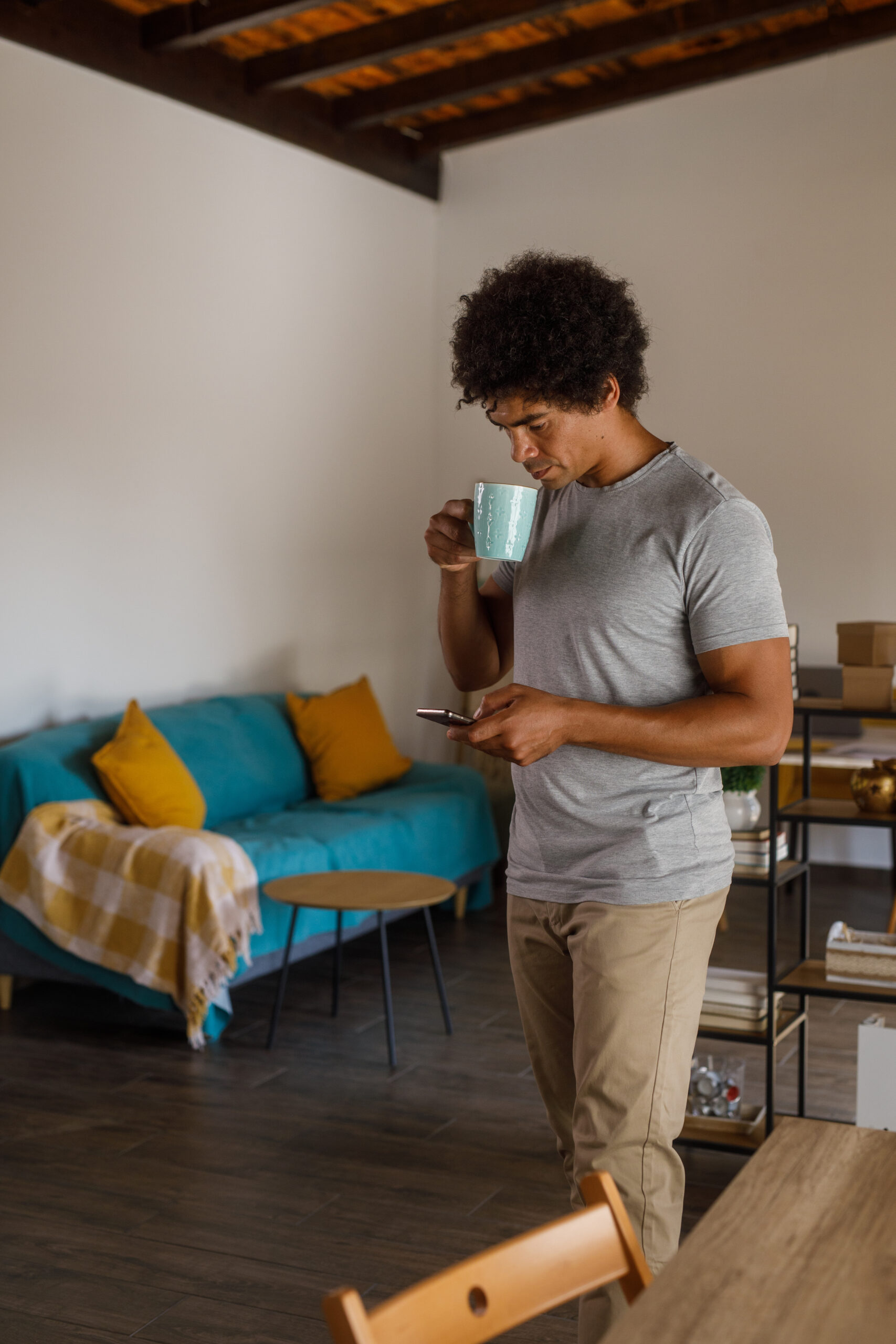 Candid shot of happy mid adult man walking through his tidy home, enjoying a cup of coffee and scrolling through funny memes online via smart phone.